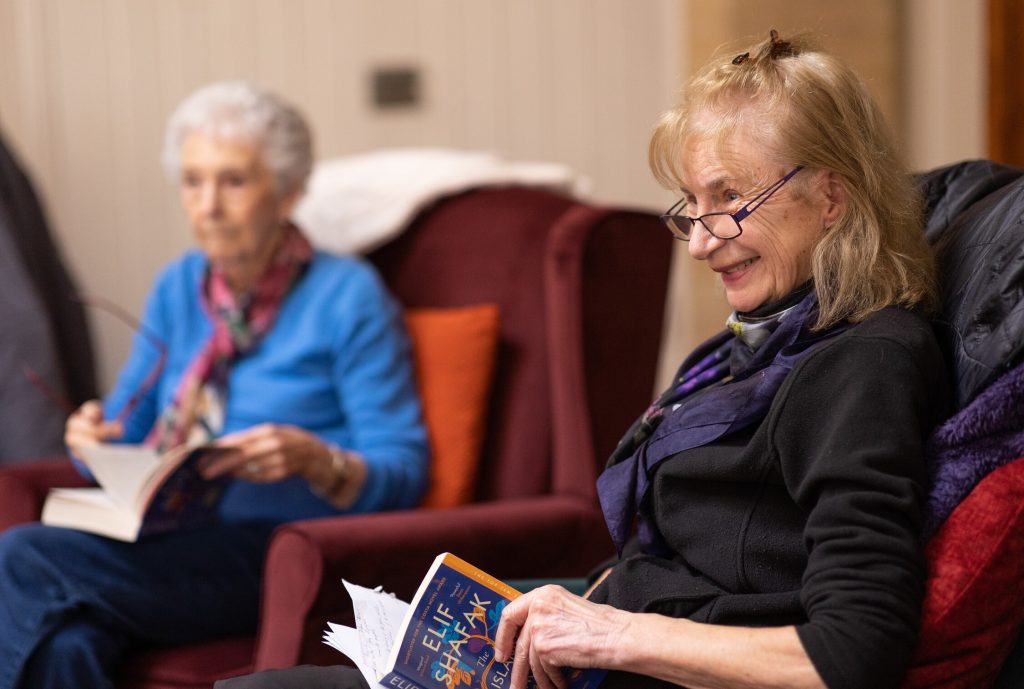 Image of a woman with a book in her hand and a woman in blue on a chair