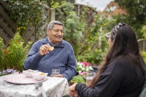 This is an image of two people talking to each other, with one eating a biscuit and the other having a drink.