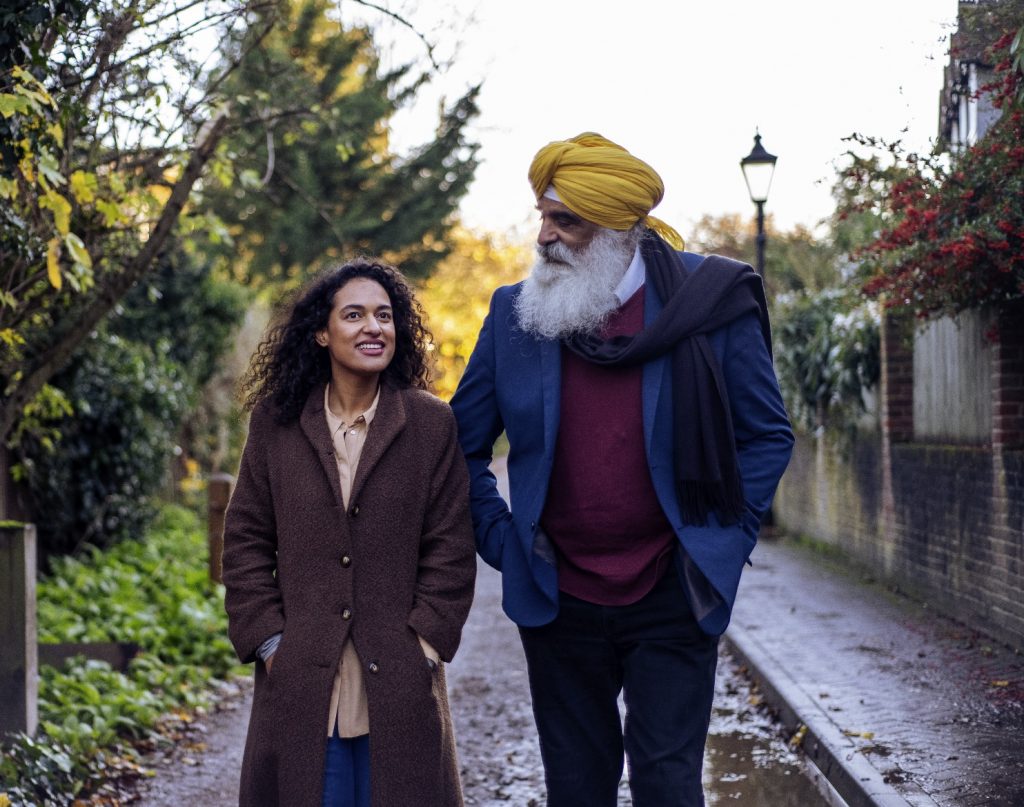 This is an image of two people walking on a street, with trees in the background.. On the left is a woman wearing a brown coat and on the right is a man wearing a yellow turban and blue blazer over a red jumper.
