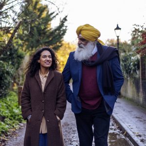 This is an image of two people walking on a street, with trees in the background.. On the left is a woman wearing a brown coat and on the right is a man wearing a yellow turban and blue blazer over a red jumper.