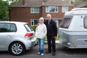 This is an image of a man and woman holding each other in front of a silver car and a caravan.
