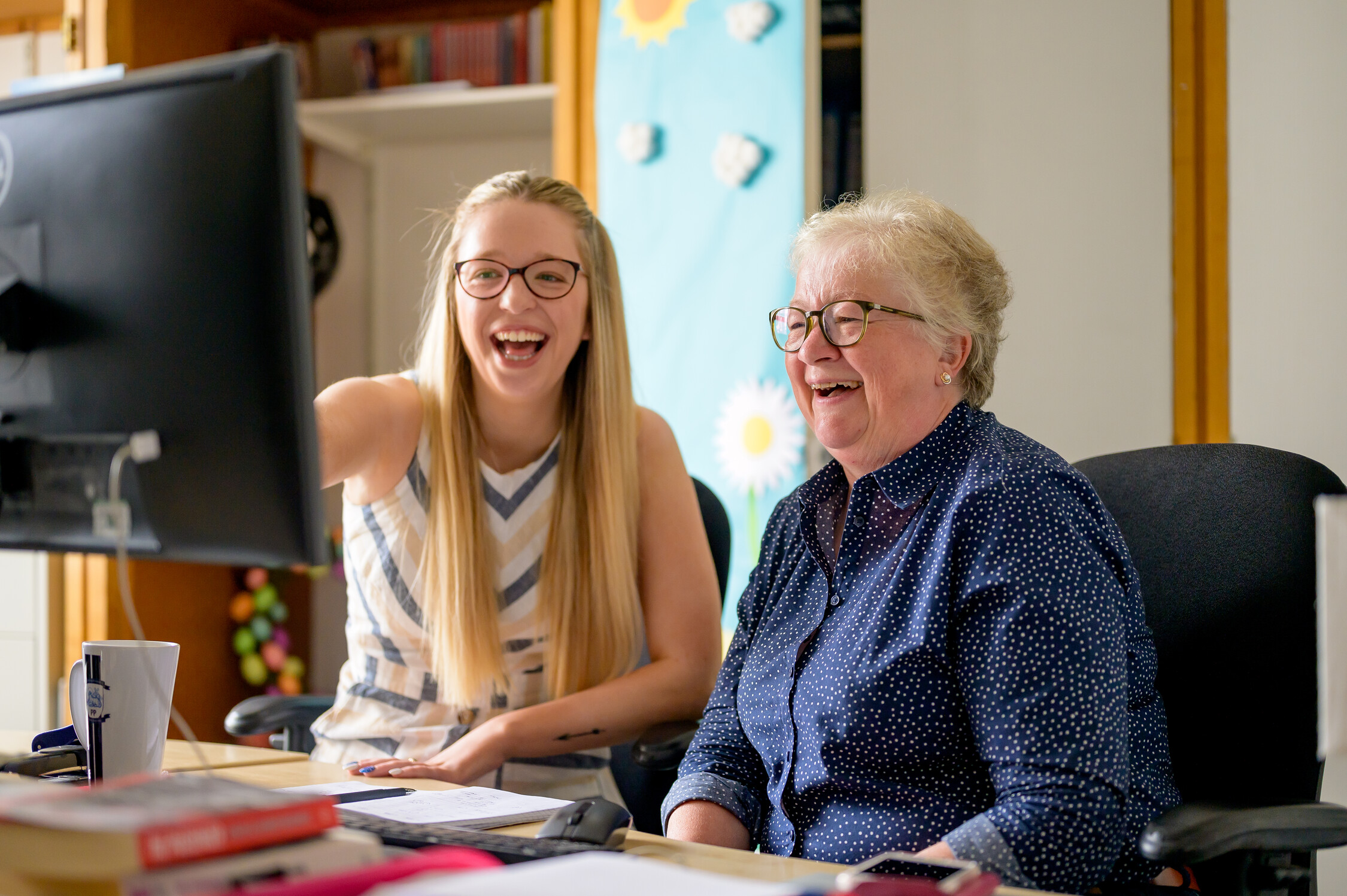 This is an image of a woman at a desk helping someone on a computer.